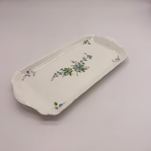 Peut inclure: Un plateau de service rectangulaire en céramique blanche avec des bords festonnés et un motif floral central composé de petites fleurs bleues et de feuilles vertes. Le plateau présente une finition lisse et brillante et un bord légèrement surélevé.