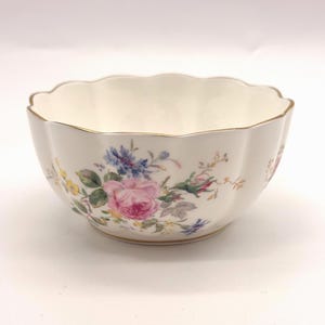 Puede incluir: Un cuenco de porcelana blanca con borde festoneado y diseño floral. El cuenco presenta rosas rosas, flores azules y amarillas y hojas verdes. El borde está adornado con oro.