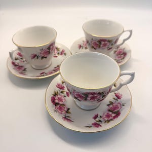 May include: Three white teacups and saucers with a pink rose pattern and gold trim. The teacups have a curved handle and sit on matching saucers. The floral design is repeated on the saucers. The set is ideal for tea parties.