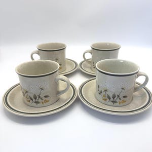 May include: Four beige ceramic coffee cups and saucers. Each cup and saucer has a black trim. The cups and saucers are decorated with a floral design in white, yellow, and brown.