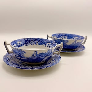 Puede incluir: Dos cuencos de sopa de cerámica azul y blanca con platillos a juego. Los cuencos tienen asas y presentan un diseño escénico con un castillo y motivos florales. Los platillos también tienen un diseño a juego. El conjunto está sobre un fondo blanco.