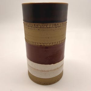 Puede incluir: Un jarrón cilíndrico de cerámica con una parte superior marrón oscuro, una banda beige con un diseño punteado, una banda granate y una franja blanca. El jarrón tiene una superficie texturizada y una paleta de colores neutros.