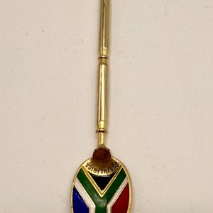 Può includere: Un cucchiaino souvenir color oro con la bandiera sudafricana sulla testa del cucchiaino. Il design della bandiera include blu, bianco, verde, rosso e nero. Il manico è lungo e cilindrico, con una parte superiore decorativa.