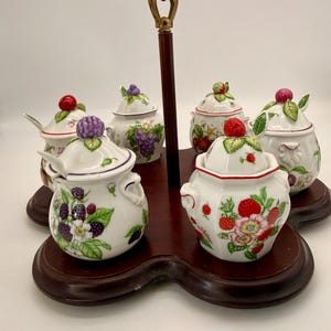 Peut inclure: Un ensemble de service à condiments composé de quatre pots en céramique blanche avec couvercles, ornés d'illustrations de fruits colorés comme des mûres, des framboises et du raisin. Les pots sont disposés sur un socle en bois foncé.