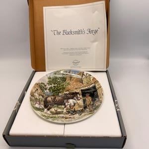 May include: A decorative plate titled "The Blacksmith's Forge" in its original box. The plate features a detailed scene of a blacksmith's shop with figures, a horse, and a building. The box is brown with a gray interior.