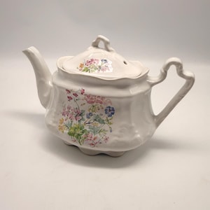 May include: A cream-coloured ceramic teapot with a floral design. The teapot has a curved handle, a spout, and a lid with a small hole. The floral pattern includes pink, blue, and yellow flowers with green leaves.