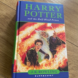 Puede incluir: Un libro de tapa dura azul con lomo verde. La portada presenta una ilustración de Harry Potter y Albus Dumbledore en una escena de fuego. El título es "Harry Potter y el Príncipe Mestizo" de J.K. Rowling. Bloomsbury.