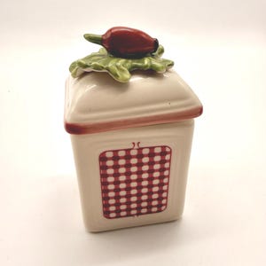 Puede incluir: Un tarro de especias de cerámica de color crema con un patrón de cuadros rojos y blancos en la parte delantera. La tapa está rematada con un pimiento rojo y hojas verdes. El tarro tiene una base de color crema con ribete rojo.