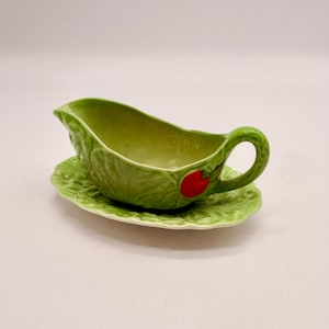 May include: A light green ceramic gravy boat with a textured cabbage leaf design and a small red apple accent on the side. The gravy boat sits on a matching oval-shaped saucer that also features a textured cabbage leaf pattern and scalloped edges.