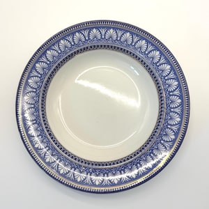 May include: A white ceramic plate with a blue and white decorative border. The border features a repeating pattern of stylized leaves and a gold trim. The plate has a wide rim and a shallow center.