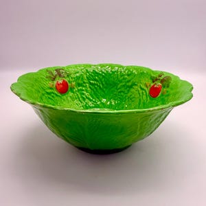 Puede incluir: Un cuenco de cerámica de color verde brillante con una superficie texturizada que recuerda a las hojas de col. El cuenco presenta un borde festoneado y dos pequeños motivos de fresas rojas en relieve con hojas verdes en el interior.