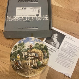 May include: A Wedgwood collector's plate featuring a painting of a horse-drawn cart with a hay bale, a family of people, and a dog. The plate is in a box with the Wedgwood logo and the words "The Artist - John L. Chapman" on the side.