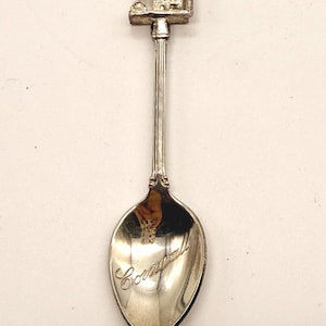 May include: Silver souvenir spoon featuring a detailed miniature building at the top, likely a landmark. The spoon's bowl is engraved with the word "Cornwall". A collectible item.