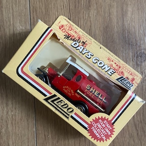 Puede incluir: Un camión de juguete rojo con una cabina blanca y un tanque de Shell Fuel Oil. El camión está en una caja amarilla y blanca con las palabras "Models of Days Gone" y "Lledo" impresas en ella.