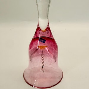 Puede incluir: Una campana de cristal transparente con un efecto degradado rosa, que va de un magenta intenso en la parte superior a un rosa más claro en la base. Un asa blanca y una cadena colgante con un acento de cristal completan el diseño. Una etiqueta azul es visible.