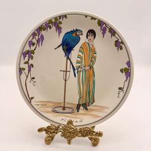 May include: A round ceramic plate featuring a colourful illustration of a person in a striped robe standing next to a blue and yellow macaw on a perch. The border is decorated with purple wisteria vines. The text N.2, Le lit is printed at the bottom.