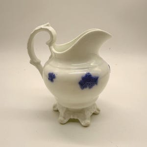 Peut inclure: Une crémière en céramique blanche avec une anse et un bec décoratifs. La crémière présente des motifs floraux bleus et repose sur une base à piédestal à trois pieds. La crémière mesure environ 15 cm de haut.