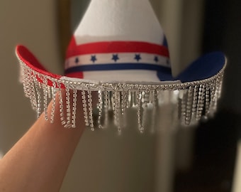 Puede incluir: Un sombrero vaquero blanco con estrellas y rayas rojas, blancas y azules. El sombrero tiene una franja plateada alrededor del borde.