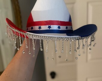 Puede incluir: Un sombrero de vaquero blanco con una banda roja, blanca y azul y estrellas. El sombrero tiene una franja de pedrería plateada.