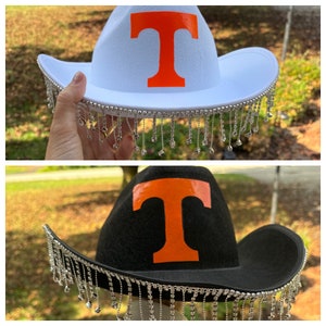 May include: Two cowboy hats with a large orange "T" on the front. The hats have a silver trim with dangling silver beads. One hat is white and the other is black.