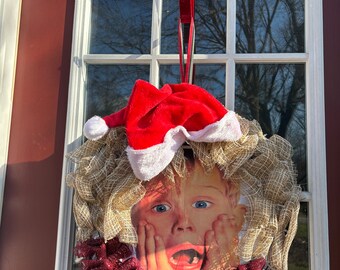 May include: A Christmas wreath with a red and white Santa hat, a burlap base, and a red and gold ribbon. The wreath features a picture of a child's face with a surprised expression.