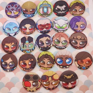 Puede incluir: Una colección de veinticuatro botones redondos y coloridos con varios diseños de personajes de dibujos animados. Los botones muestran una variedad de caras con ojos grandes, en tonos de azul, verde, marrón y morado. Los botones están dispuestos sobre una superficie estampada.