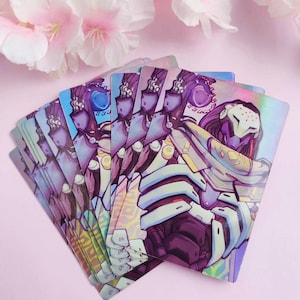 May include: A stack of holographic cards featuring a purple and white illustration of a robot character with a helmet and a scarf. The character is holding a weapon. The cards have a rainbow iridescent finish.