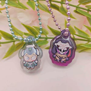 May include: Two pendant necklaces with cartoon character designs. One pendant is light blue and the other is purple. Each pendant is attached to a beaded chain. The background features green leaves and a pink and white surface.