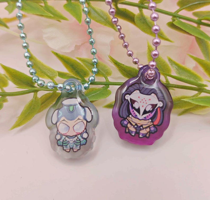 May include: Two pendant necklaces with cartoon character designs. One pendant is light blue and the other is purple. Each pendant is attached to a beaded chain. The background features green leaves and a pink and white surface.
