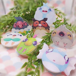 Puede incluir: Colección de siete colgantes acrílicos de personajes de dibujos animados coloridos. Cada colgante presenta un diseño único con varios colores, incluyendo azul, verde, rosa y marrón. Los colgantes están dispuestos con follaje verde en el fondo.