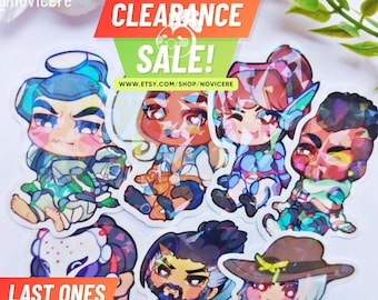 Puede incluir: Una colección de siete pegatinas holográficas con personajes estilo chibi de diversos colores de pelo, atuendos y expresiones. Las pegatinas tienen un acabado prismático reflectante y están agrupadas. El texto incluye CLEARANCE SALE!