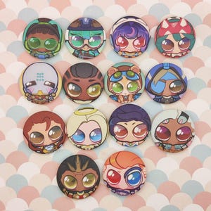 Puede incluir: Una colección de botones redondos y coloridos con diseños de personajes de dibujos animados con ojos grandes y varios atuendos. Los botones tienen un acabado brillante y están dispuestos sobre una superficie estampada en rosa y blanco. Cada botón muestra un personaje único.
