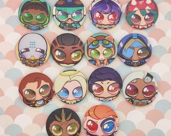 May include: A collection of colorful, round button pins featuring cartoon character designs with large eyes and various outfits. The pins have a glossy finish and are arranged on a pink and white patterned surface. Each pin showcases a unique character.
