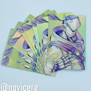 May include: A stack of iridescent cards featuring a character from the video game Overwatch. The character is wearing a white helmet and a purple and white hooded cloak. The character's hands are visible, and they are holding a weapon. The cards have a holographic finish.