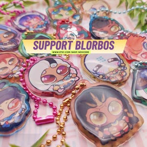 Op de afbeelding: Een verzameling acrylbedels met diverse karakterillustraties in chibi-stijl, elk bevestigd aan een gekleurde metalen kogelketting. Een centrale gele banner toont de tekst SUPPORT BLORBOS en WWW.ETSY.COM/SHOP/NOVICERE in het paars.