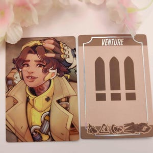 May include: Two trading cards featuring a character illustration and a card back design. The character card shows a person in a yellow and tan outfit. The card back has the word "VENTURE" and three exclamation marks.