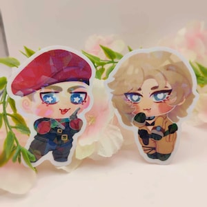 Może przedstawiać: Dwie naklejki z postaciami w stylu anime. Jedna postać nosi czerwony beret i trzyma broń, druga ma blond włosy i mikrofon. Obie mają źrenice w kształcie gwiazd. Naklejki mają efekt holograficzny.