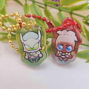 May include: Two acrylic keychains with cartoon character designs. One keychain has a gold ball chain and features a character with a white helmet and green accents. The other has a red ball chain and a character with blue eyes.