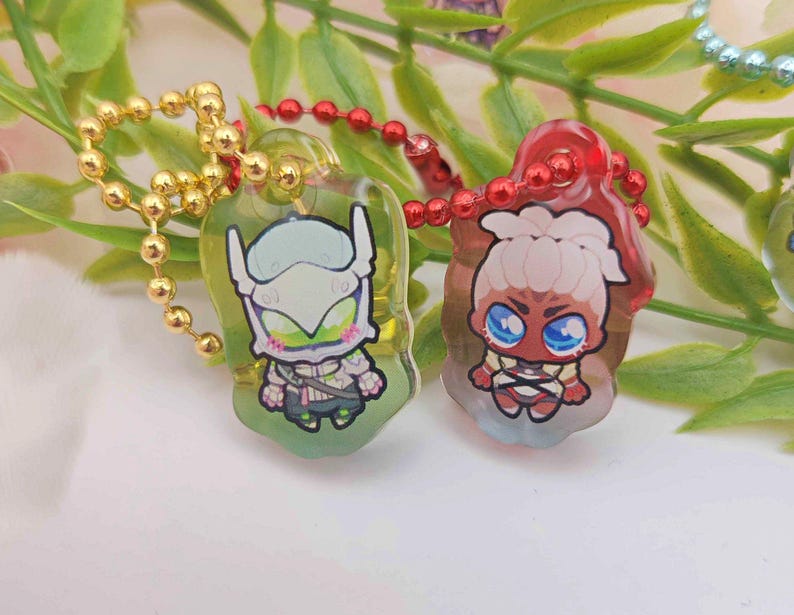May include: Two acrylic keychains with cartoon character designs. One keychain has a gold ball chain and features a character with a white helmet and green accents. The other has a red ball chain and a character with blue eyes.