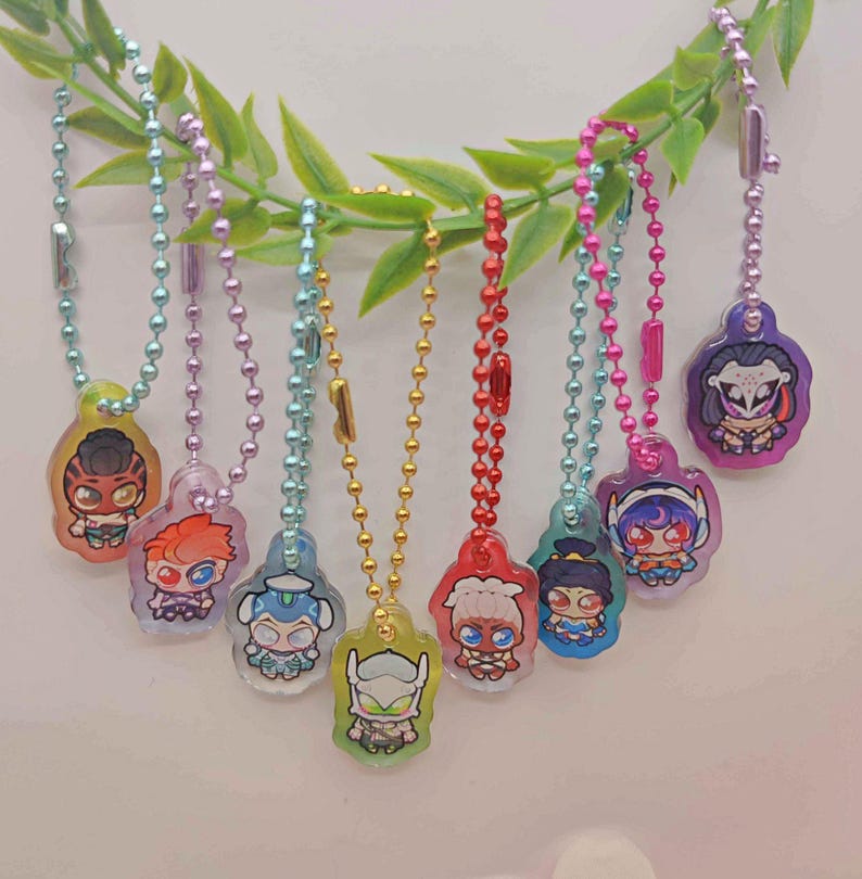 May include: A collection of eight colorful character keychains hanging from a green leafy branch. Each keychain features a different cartoon character design, attached to a ball chain. The keychains are in various colors, including yellow, blue, red, and purple.