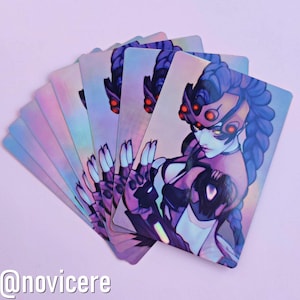 May include: A stack of iridescent holographic cards featuring a purple-haired anime character with red eyes and a black and white outfit. The character is wearing a mask with red accents. The cards have a holographic finish and are printed with a purple and blue gradient.