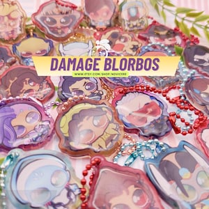 Puede incluir: Una colección de dijes acrílicos coloridos con varias ilustraciones de personajes de dibujos animados, cada uno unido a una pequeña cadena de bolas de metal. Una pancarta amarilla muestra el texto DAMAGE BLORBOS y WWW.ETSY.COM/SHOP/NOVICERE.