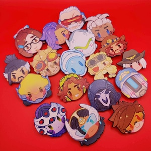 Könnte beinhalten: Eine Sammlung farbenfroher, cartoonartiger Charaktergesichtsanstecker auf rotem Hintergrund. Die Anstecker zeigen verschiedene Charaktere mit unterschiedlichen Frisuren, Accessoires und Gesichtsausdrücken. Die Anstecker sind rund und scheinen aus glänzendem Material zu bestehen.