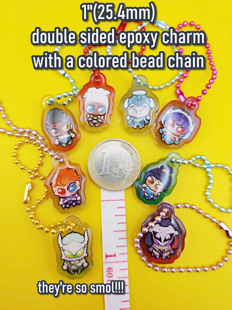 May include: A collection of double-sided epoxy charms, each featuring a unique cartoon character design. The charms are approximately 1 inch (25.4mm) in size and come with a colored bead chain. Text on the image reads "they're so smol!!!".