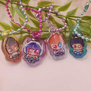 May include: Four colorful pendant necklaces featuring cartoon characters. Each pendant is a different color and design, with a silver ball chain and a pink or blue beaded chain. The pendants are displayed against a backdrop of green leaves and pink flowers.