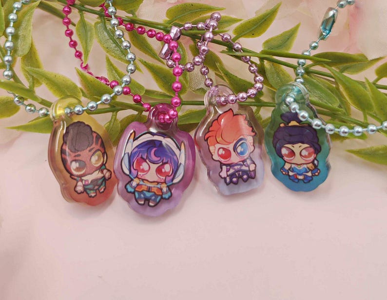 May include: Four colorful pendant necklaces featuring cartoon characters. Each pendant is a different color and design, with a silver ball chain and a pink or blue beaded chain. The pendants are displayed against a backdrop of green leaves and pink flowers.