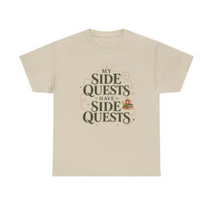 May include: A beige short-sleeved t-shirt featuring dark green text that reads "MY SIDE QUESTS HAVE SIDE QUESTS" in a decorative serif font. The design includes small star accents, a dotted path graphic, and a stack of books with a coffee mug.