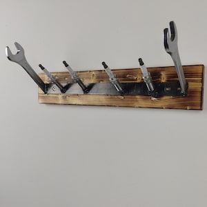 Könnte beinhalten: Ein Wandregal aus Holz mit sechs Haken, ausgestattet mit fünf Zündkerzen und einem Schraubenschlüssel. Das Regal ist dunkelbraun gebeizt und hat einen rustikalen Look.