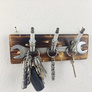Könnte beinhalten: Ein Schlüsselhalter aus Holz mit drei Zündkerzen und zwei Schraubenschlüsseln. Der Halter hat eine gebrannte Holzoptik und ist an einer weißen Wand montiert. Ein Schlüsselsatz hängt am Halter.