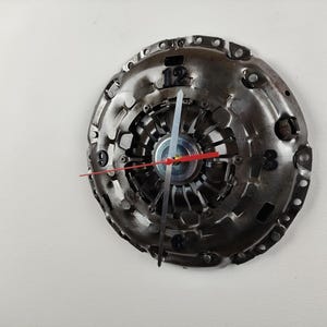 Könnte beinhalten: Eine Uhr aus einem wiederverwendeten Kupplungsplatte eines Autos. Das Zifferblatt der Uhr ist aus Metall und hat schwarze Zahlen. Die Zeiger sind silber und rot.